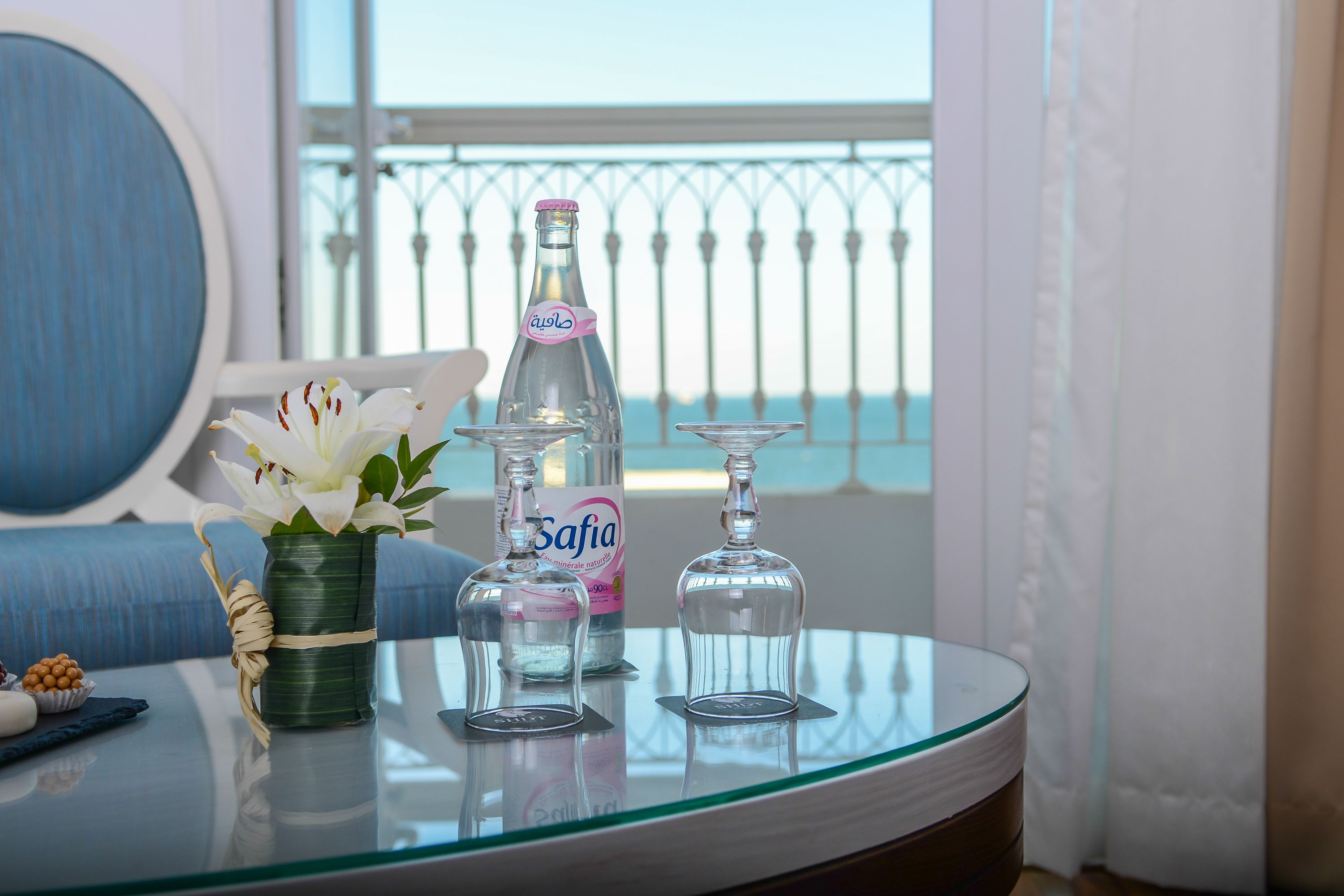 A glass table with a bottle of Safia water, a vase with white flowers, and two empty wine glasses.