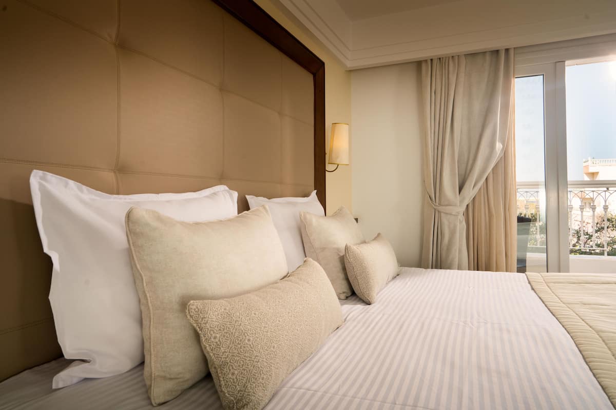 Habitación de hotel con una cama grande, varios almohadones, cabecera, lámpara de pared y vistas al exterior a través de una ventana con cortinas.