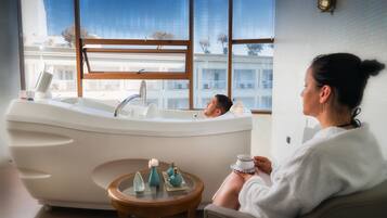 A modern bathroom with a freestanding bathtub, a small round table with decorative items, and a view of a building through large windows.