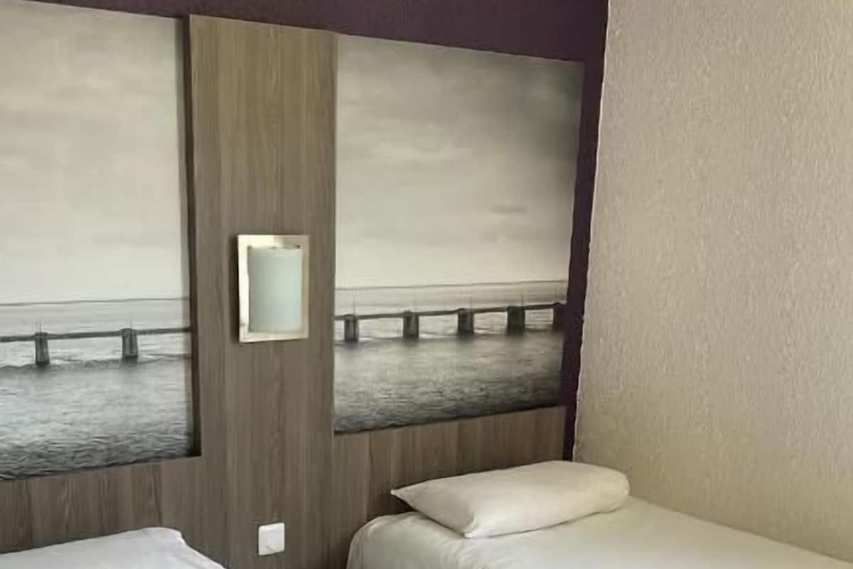 Quarto de hotel com duas camas de solteiro, uma mesa de cabeceira e uma luminária fixada na parede.