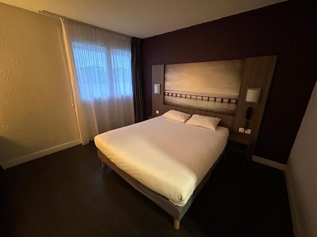 Une chambre d’hôtel avec un grand lit, une tête de lit en bois, une table de chevet avec une lampe et une fenêtre avec des rideaux.