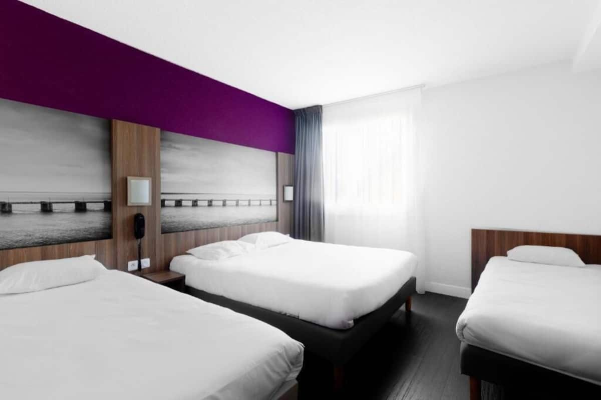 Quarto de hotel com três camas, uma janela grande com cortinas e vista para o mar, e uma parede de destaque roxa.