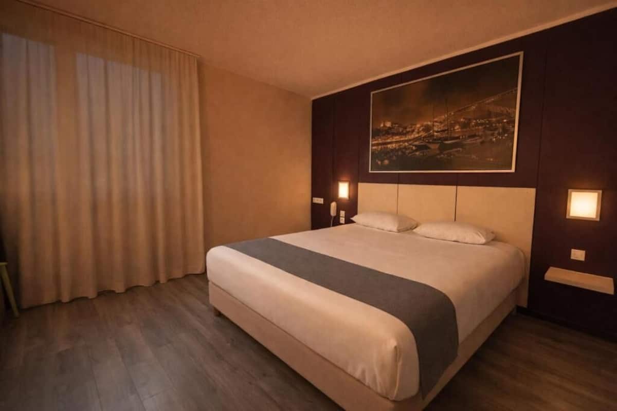 Quarto de hotel com uma cama grande, um quadro de paisagem urbana na parede e duas luminárias de cabeceira.