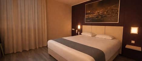 A hotel room with a large bed, a cityscape painting on the wall, and two bedside lamps.