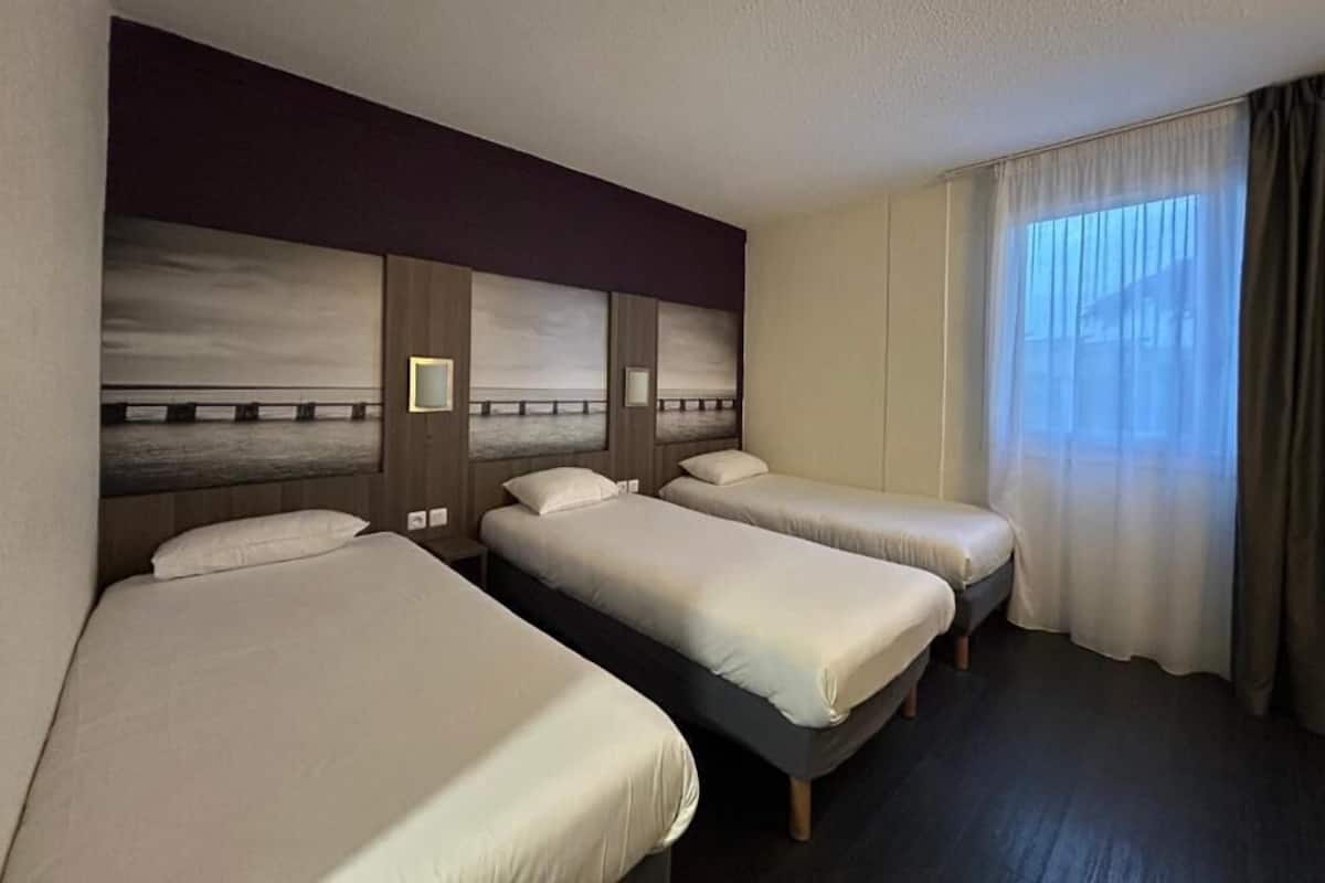 Quarto de hotel com três camas de solteiro, uma janela grande com cortinas e um design moderno para a cabeceira.