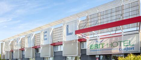 Il s'agit d'un hôtel au design moderne, avec une enseigne bien visible indiquant qu'il fait partie du groupe Brit Hotel. Plusieurs voitures sont garées devant, et le bâtiment possède un espace extérieur couvert agrémenté de plantes.