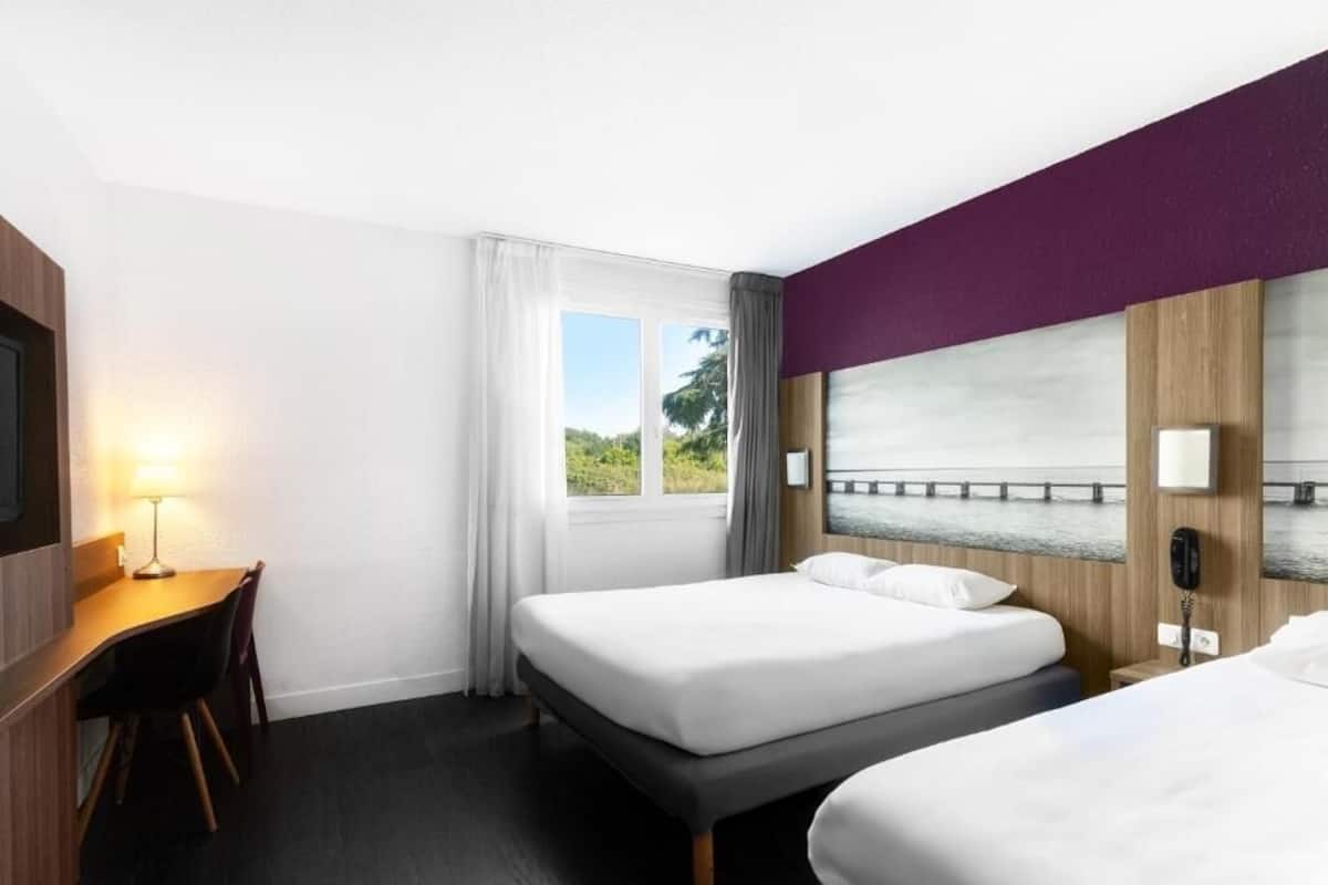 Quarto de hotel com duas camas, uma escrivaninha, uma cadeira, uma janela com cortinas e vista para áreas verdes.