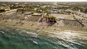 An aerial view of a beach with numerous umbrellas and people, a building with a red roof, and a clear sky. - Tui Magic Life Penelope Beach (Djerba Midun)