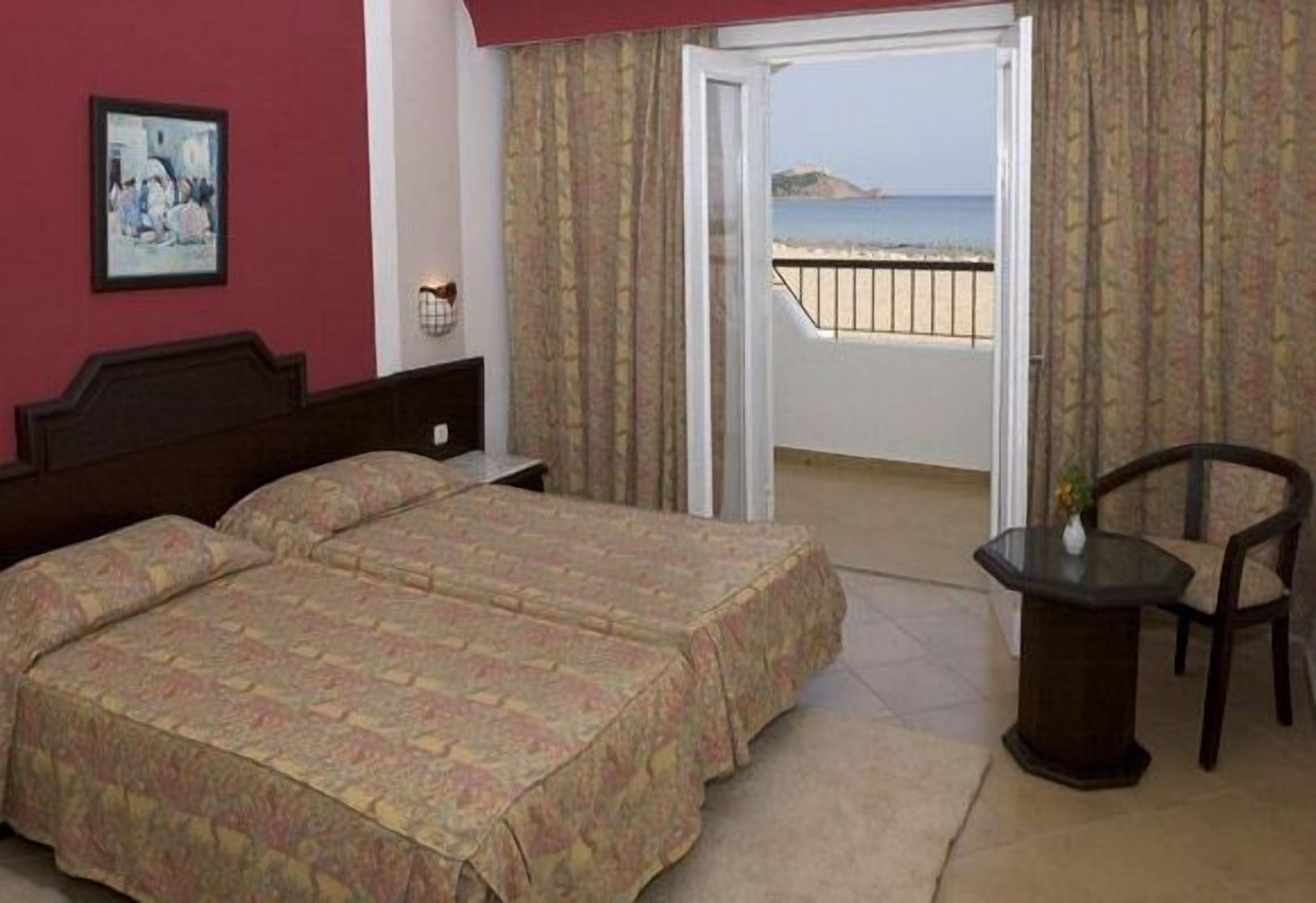A hotel room with a bed, a chair, a small table, and a view of the beach.