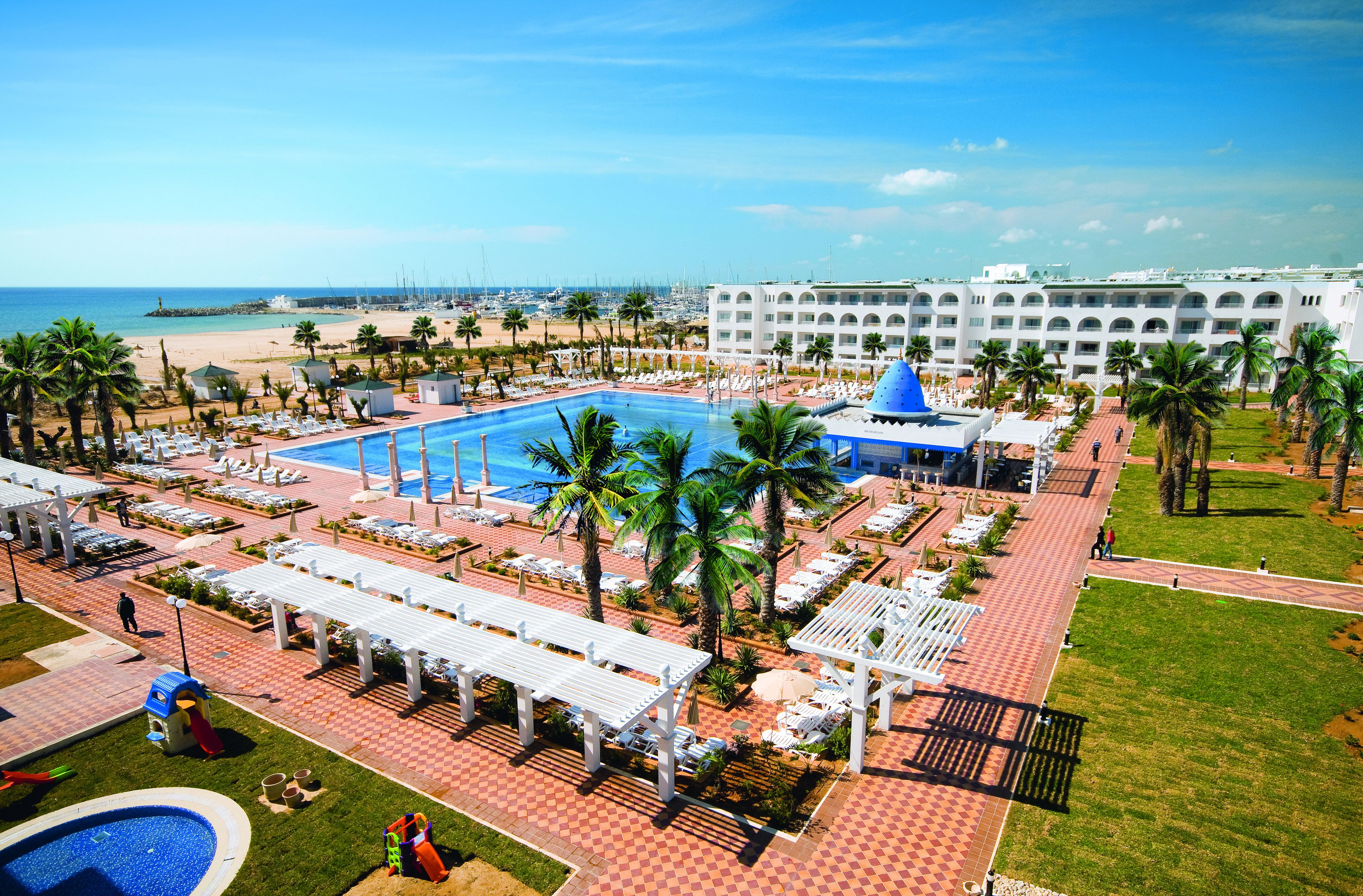 Un resort con piscina centrale, lettini, palme e vista sulla spiaggia.