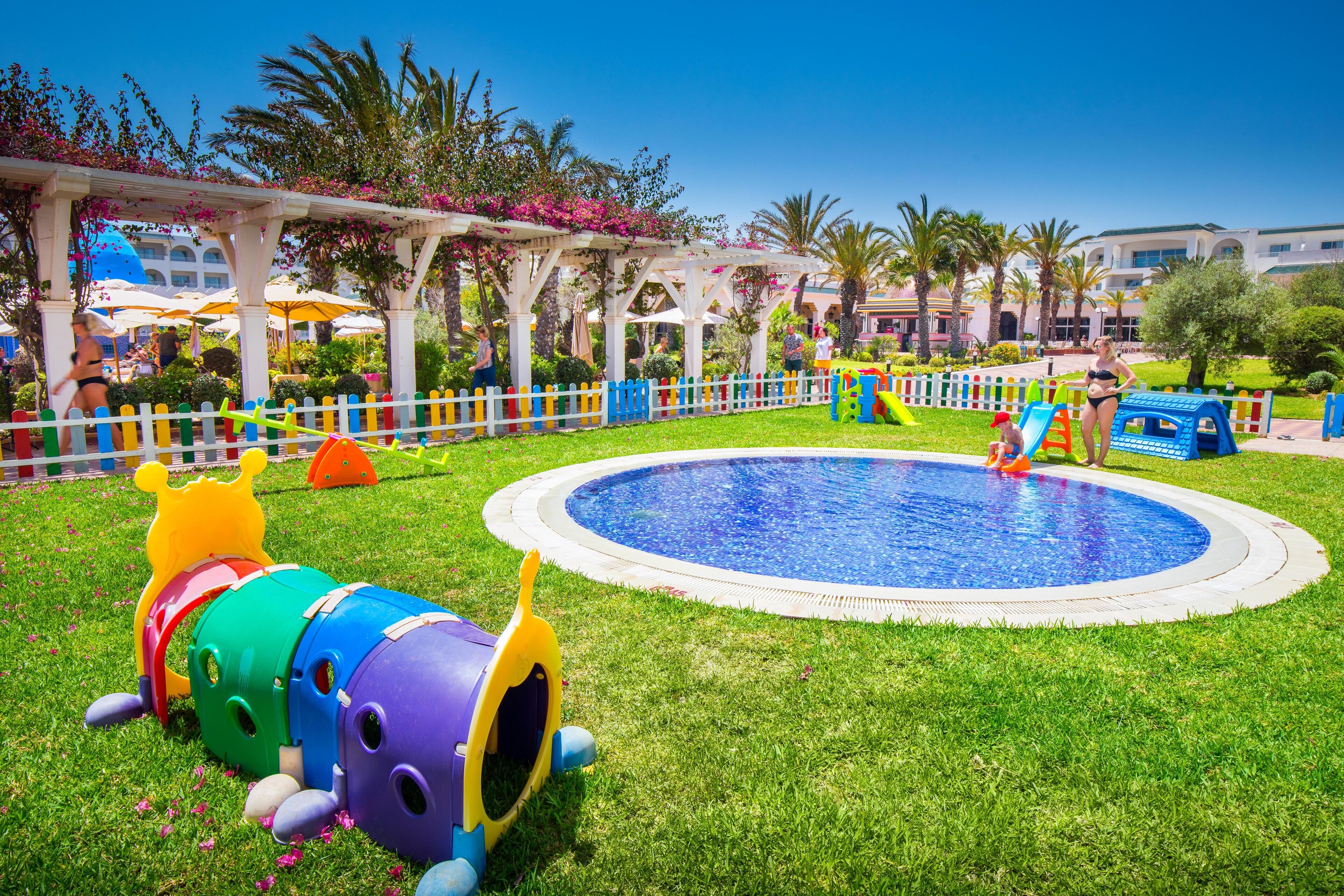 A pool area with a colorful play structure, a white fence, and palm trees.