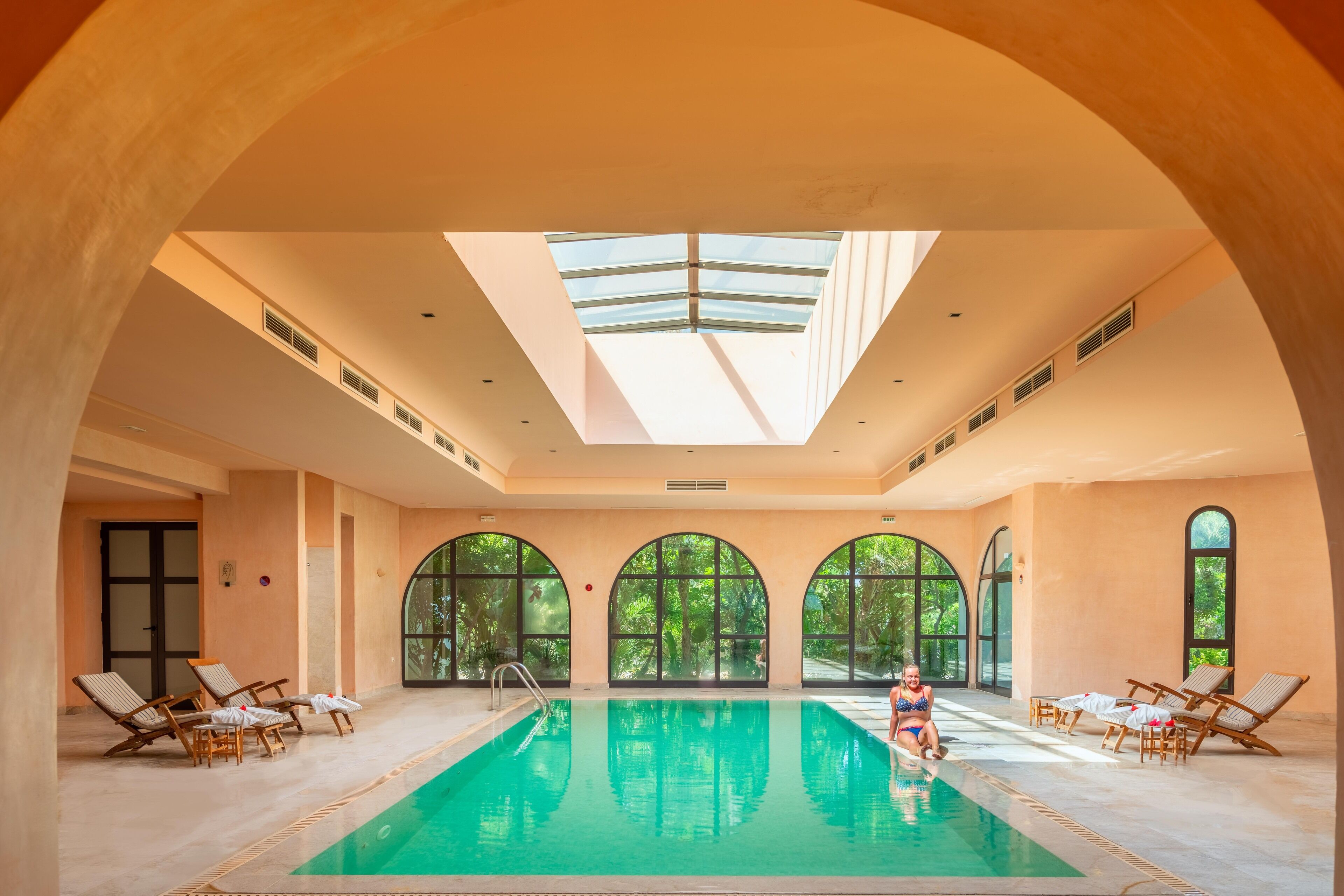 An indoor swimming pool with clear glass ceilings, multiple windows, and lounge chairs.