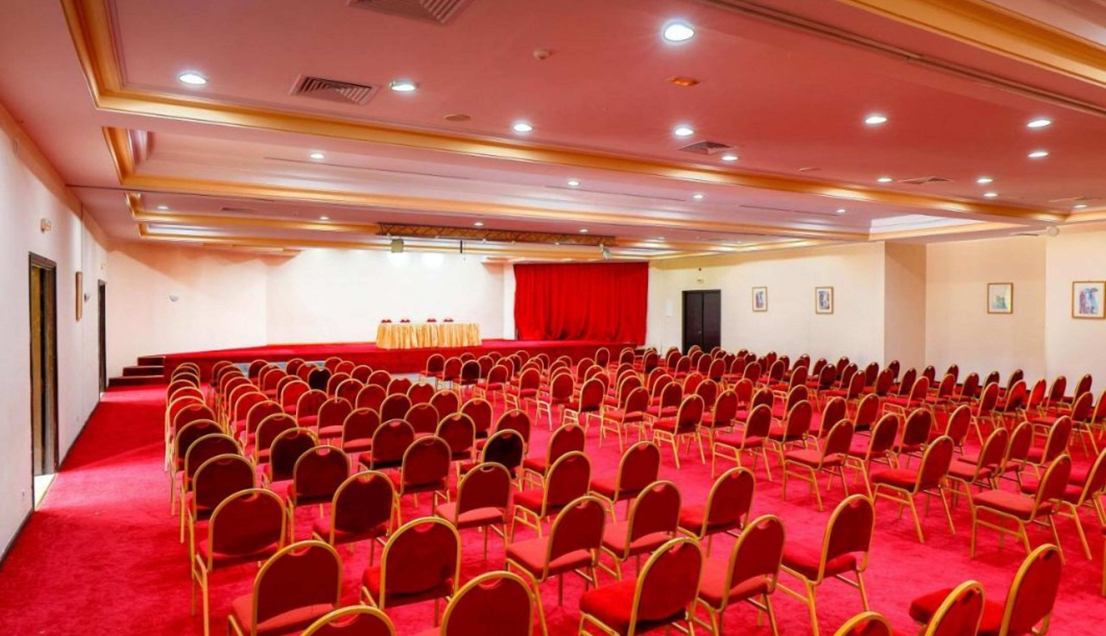 Een congreszaal met rijen rode stoelen, een podium met een rode achtergrond en een vloerbedekking.
