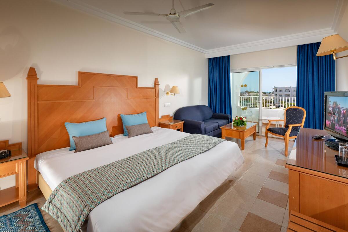 A hotel room with a large bed, a desk, a chair, a TV, and a balcony with a view.
