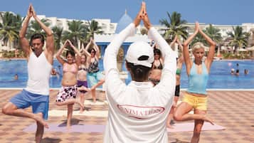 Un grupo de personas practicando yoga sobre una esterilla a cuadros junto a una piscina, con palmeras y un cielo despejado al fondo.