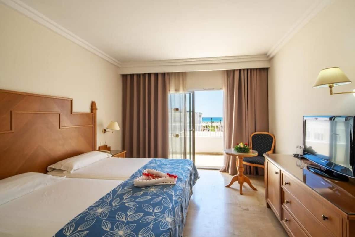 A hotel room with two beds, a television, a desk, and a view of the sea.