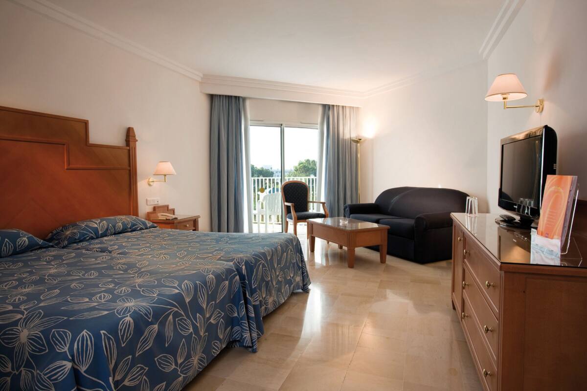 A hotel room with a large bed, a sofa, a TV, a desk, and a balcony with curtains.