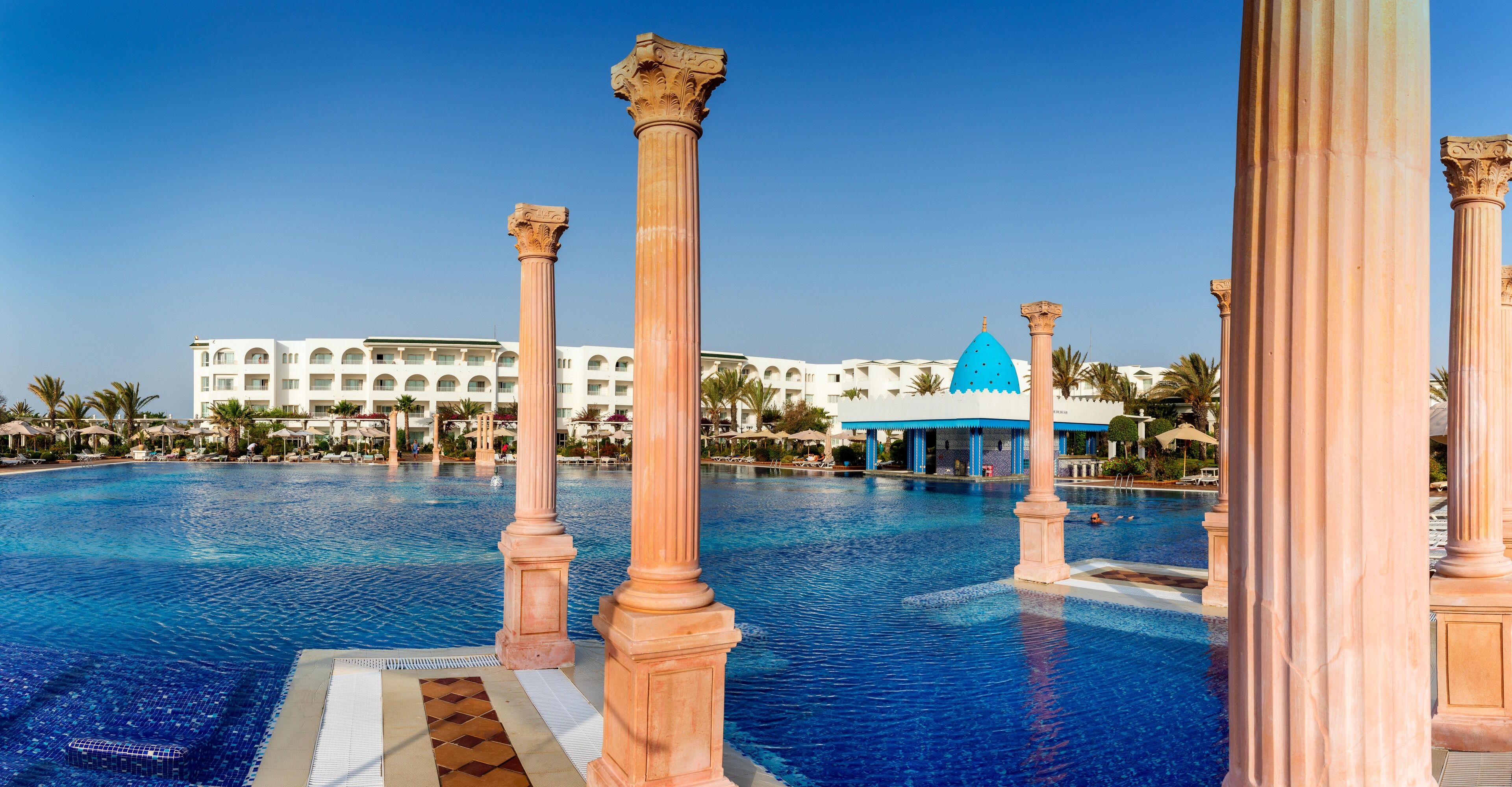 A swimming pool with classical columns and a hotel in the background.