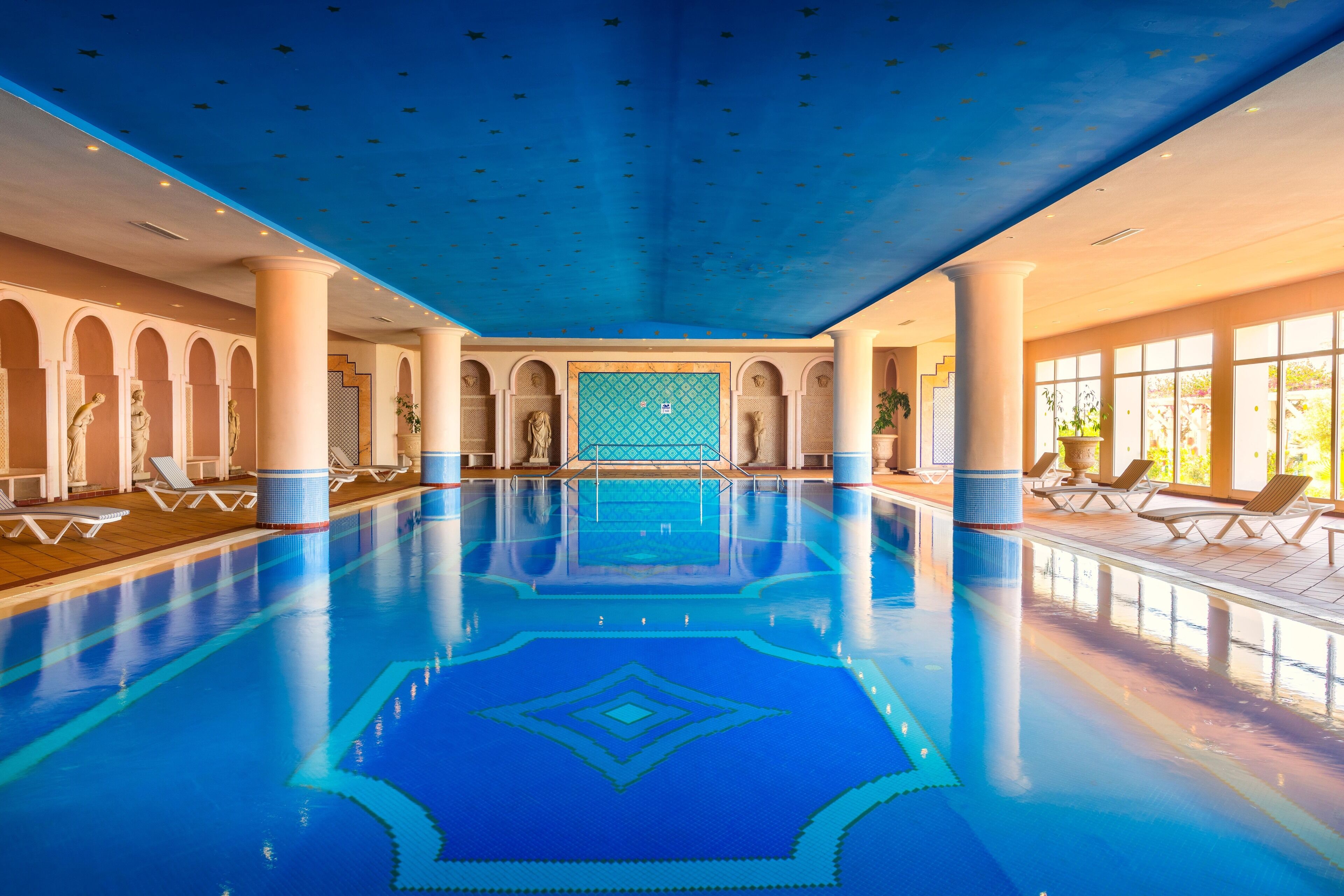 An indoor swimming pool with clear blue water, white columns, and sunlit areas with lounge chairs.