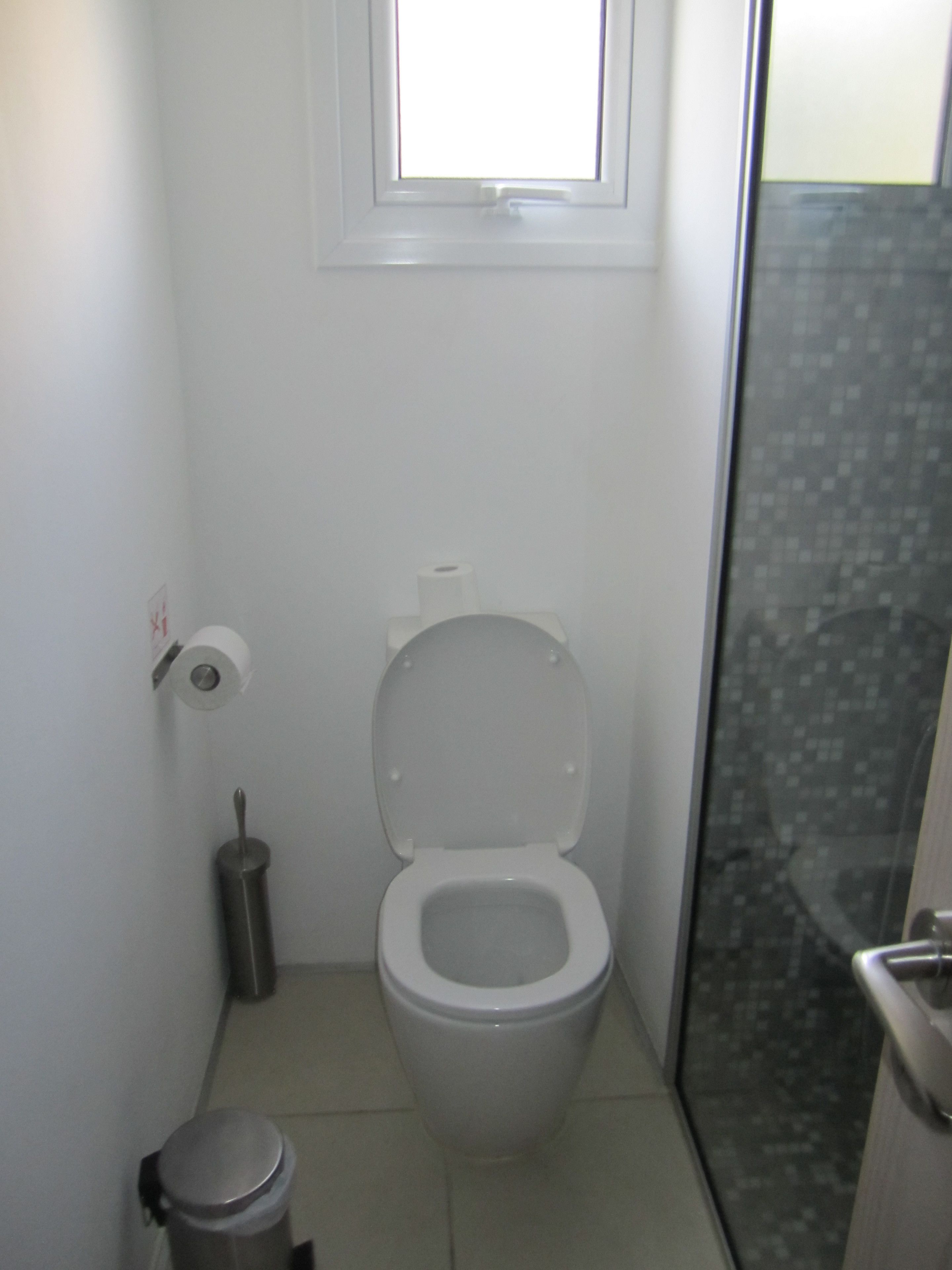 A small, white bathroom with a toilet, a trash can, and a window.
