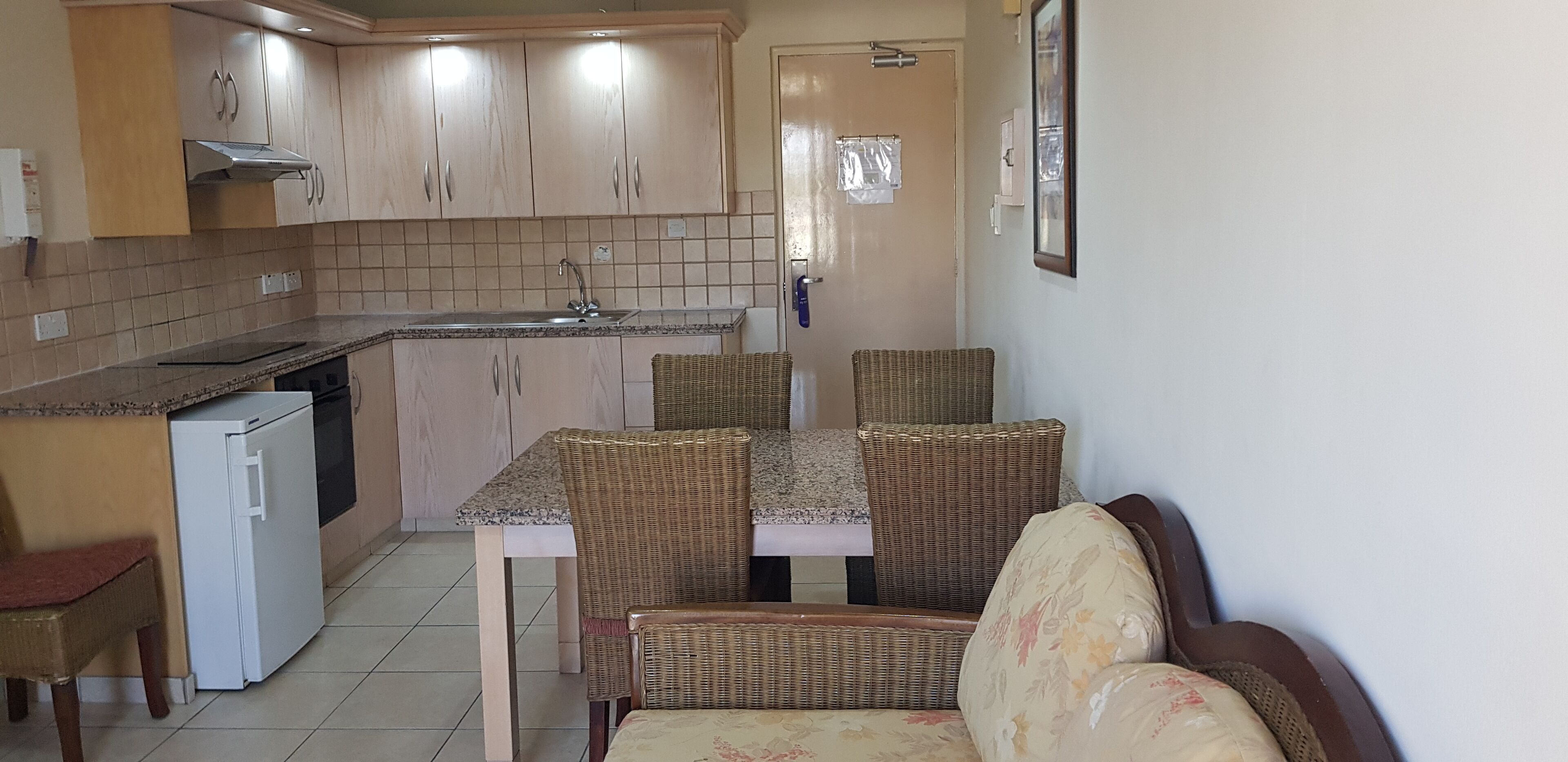 A compact kitchen with a dining area, including a refrigerator, a sink, and wicker chairs.