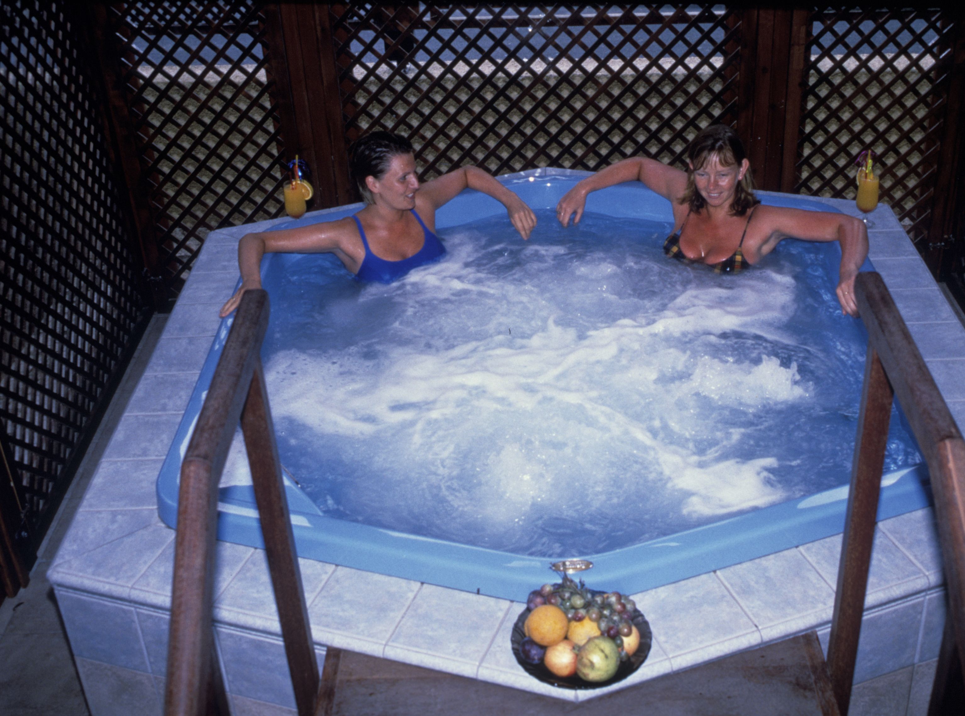 A hot tub with two people, surrounded by a wooden fence and a tiled floor.
