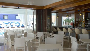 A restaurant with white wicker furniture, a bar area with bottles, and a view of a blue building outside. - Senator Hotel Apartments - Adults Only (Ayia Napa)