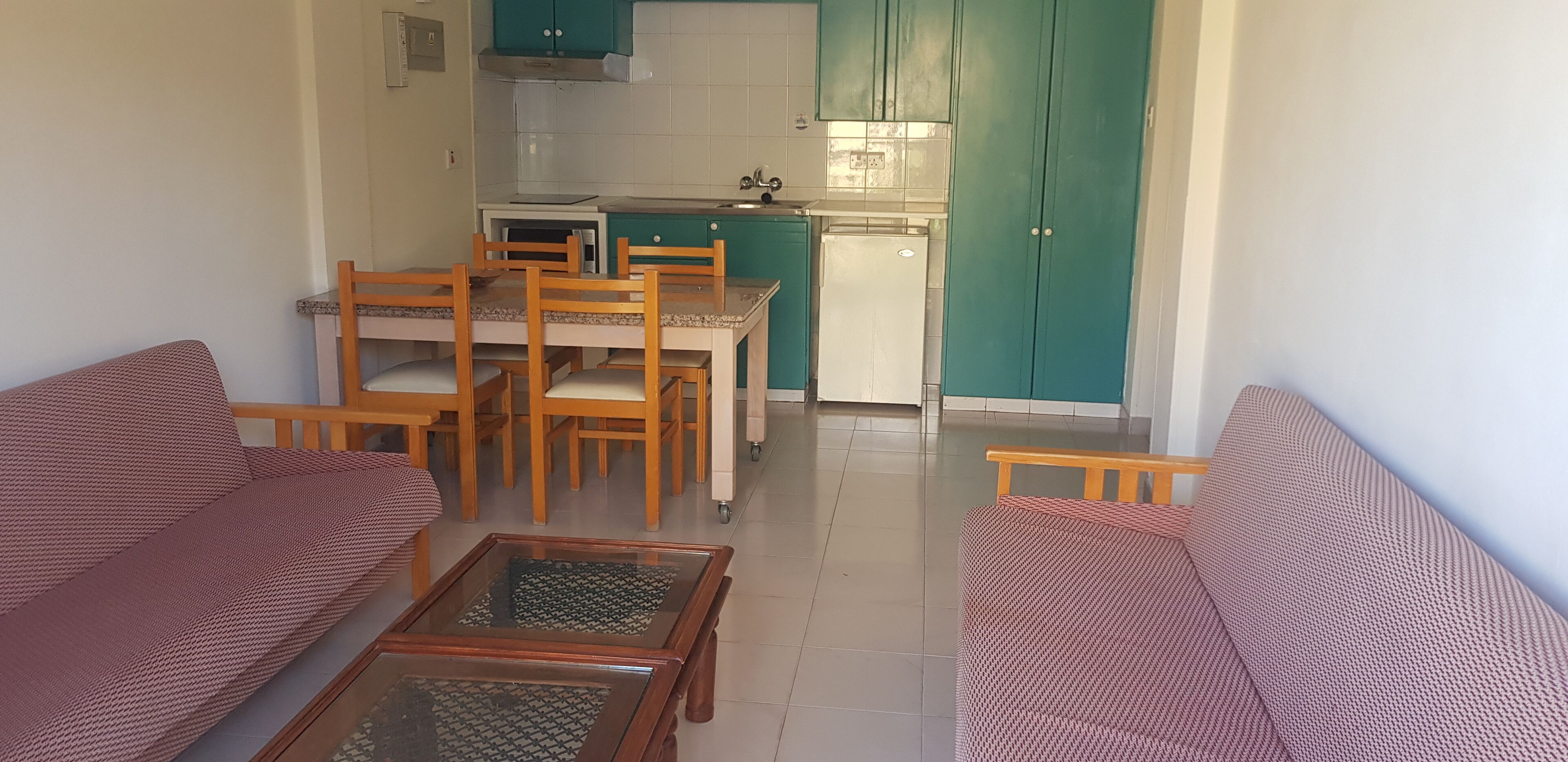 A living room with two sofas and a coffee table, a dining area with a table and chairs, and a kitchen with teal cabinets and white appliances.