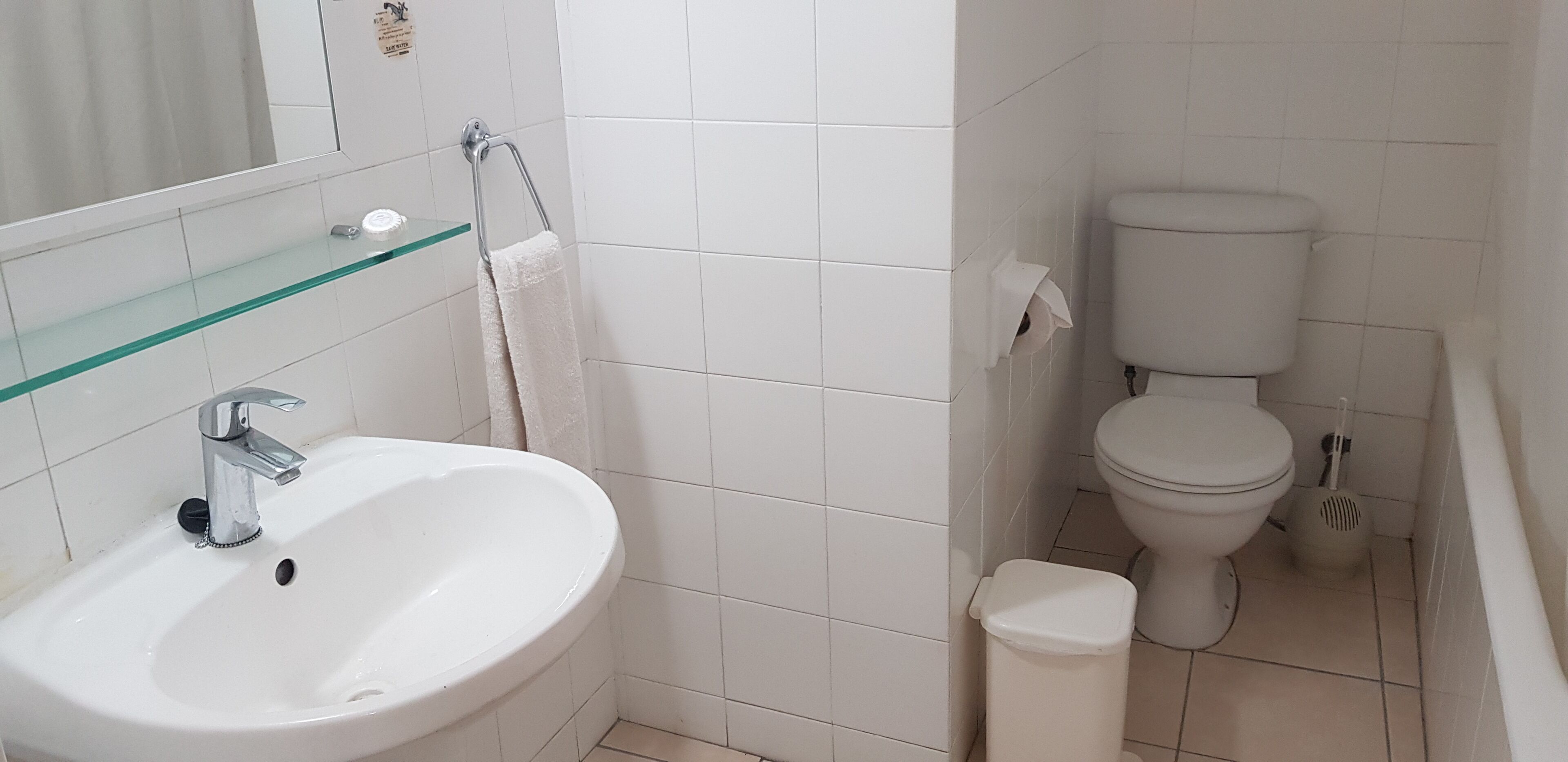 A bathroom with a white sink, a glass shelf, a toilet, and a trash bin.