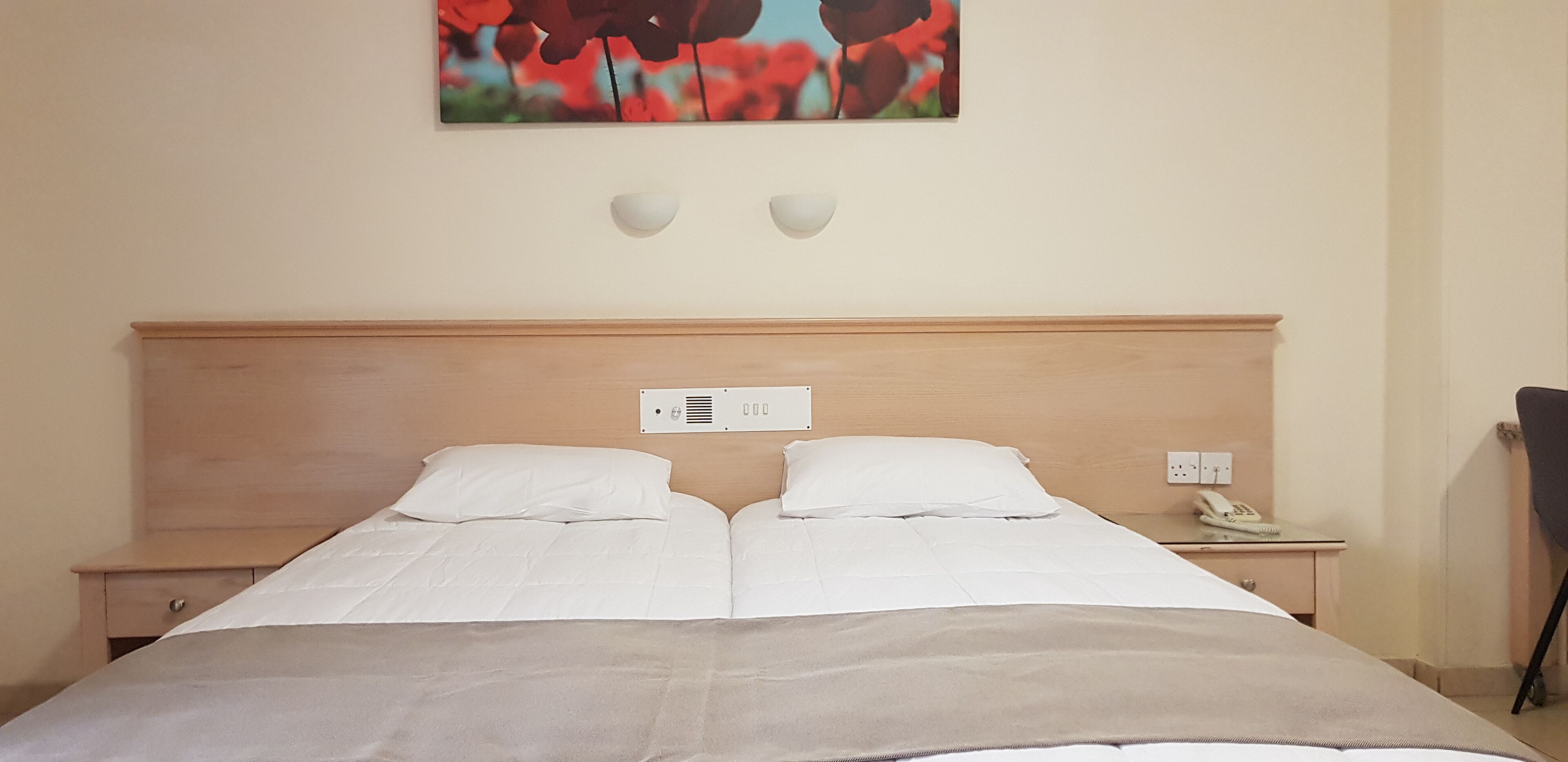 Un lit double avec une literie blanche, une tête de lit en bois, deux tables de chevet, un luminaire fixé au mur et un tableau représentant des fleurs rouges.