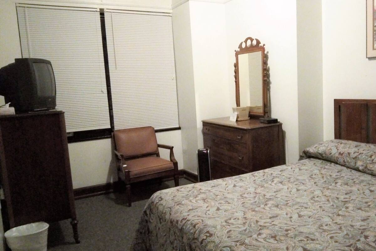 Une chambre à coucher comprenant un lit, une commode, une chaise, un téléviseur posé sur un meuble et une fenêtre avec des stores.