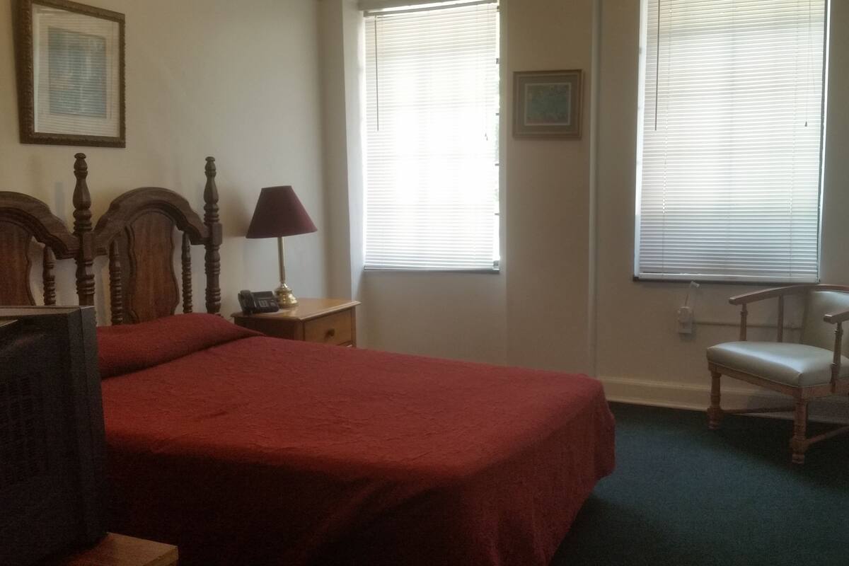 Une chambre à coucher comprenant un lit, une chaise, une table de chevet, une lampe et deux fenêtres équipées de stores.