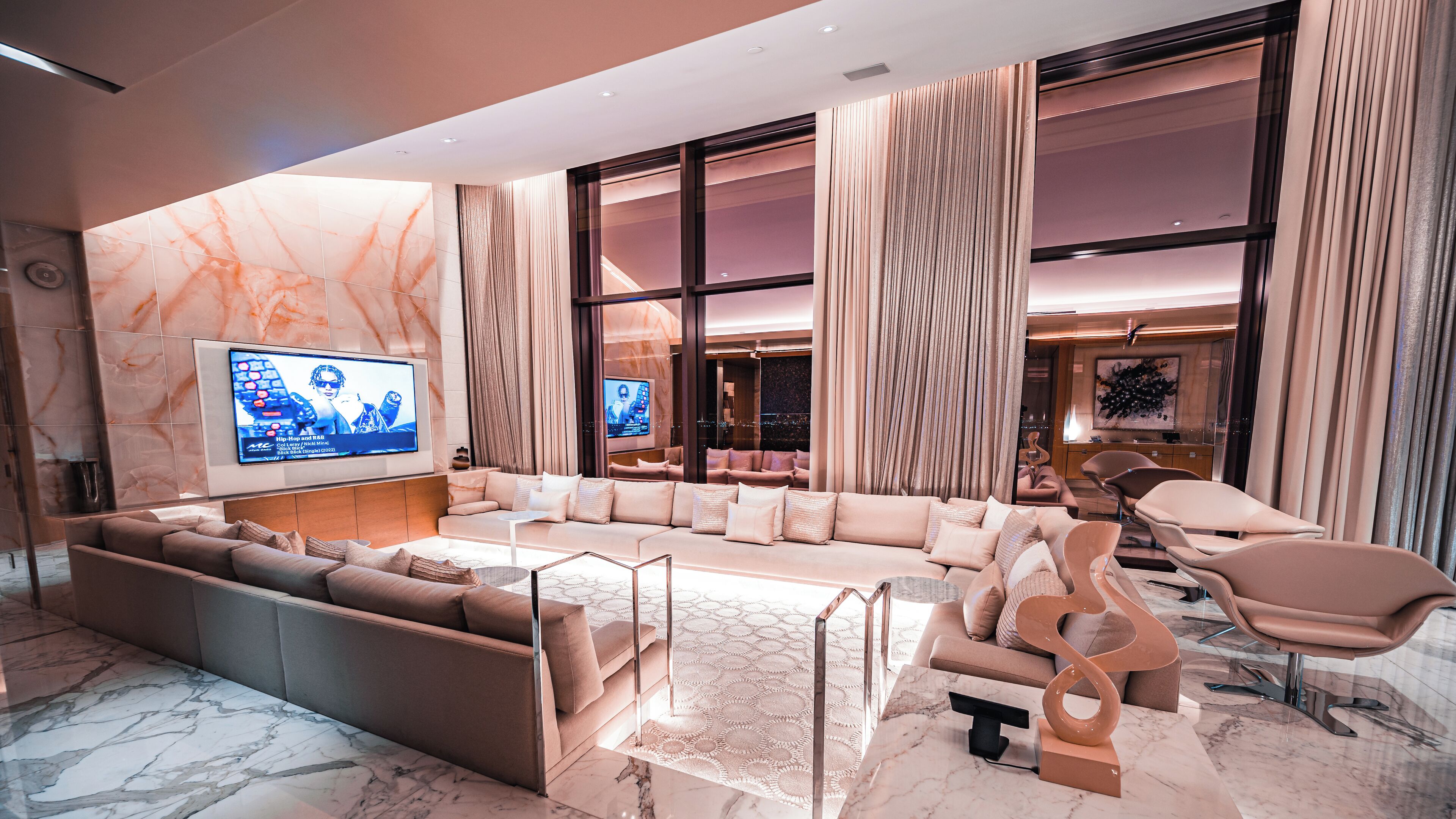 A modern living room with a large sectional sofa, a flat-screen TV mounted on the wall, and a large window with curtains.
