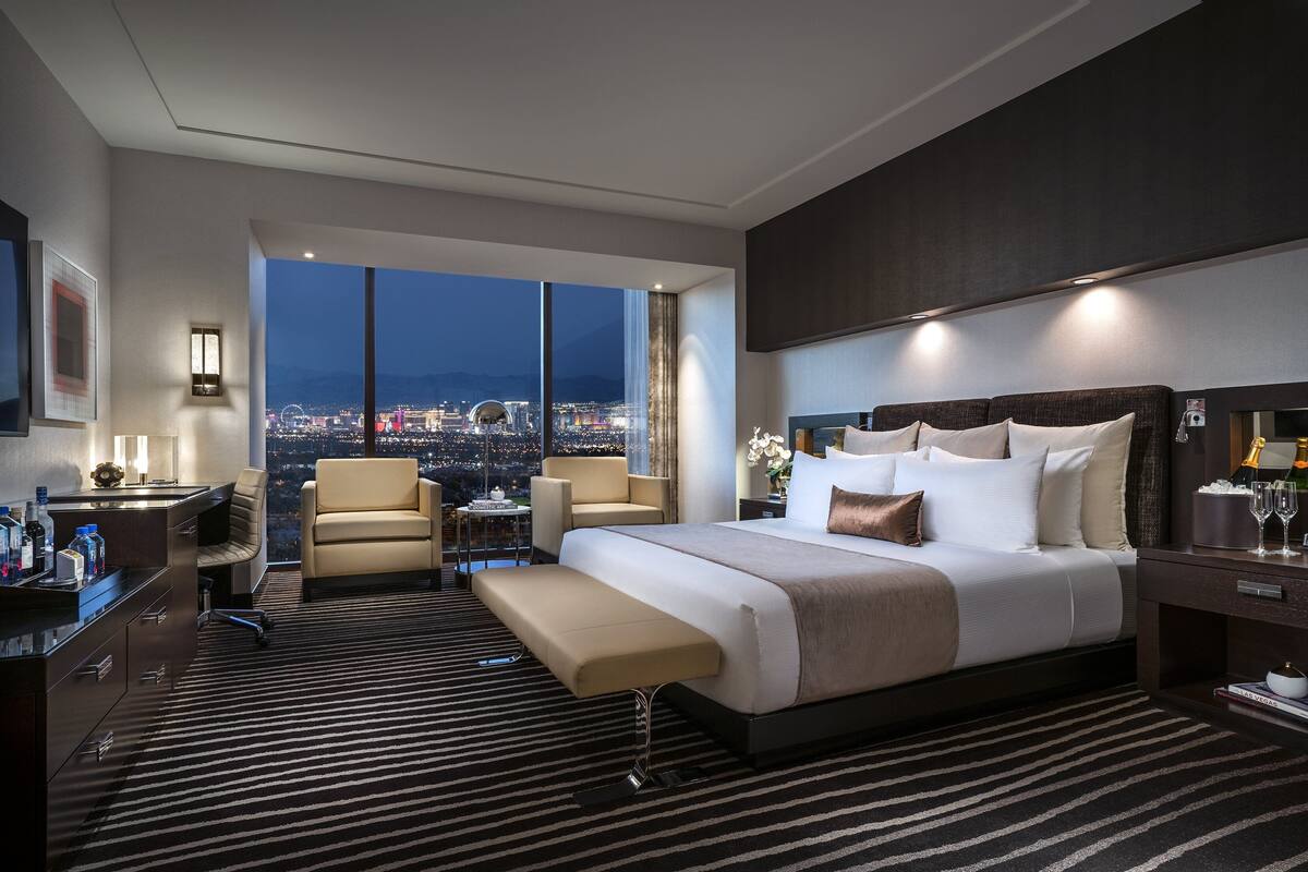 Een ruime hotelkamer met een groot bed, een zithoek, een bureau en uitzicht op een stadsbeeld bij nacht.