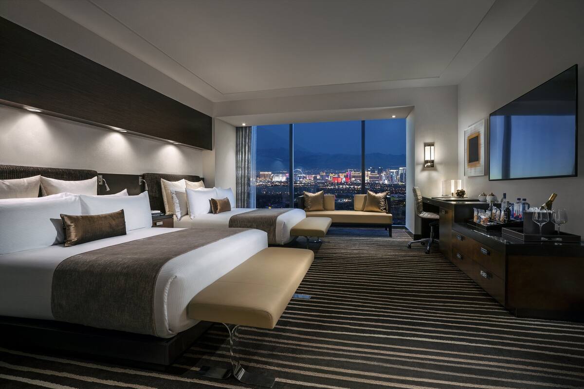 Een hotelkamer met twee bedden, een bank, een bureau en een groot raam met uitzicht op de stad.