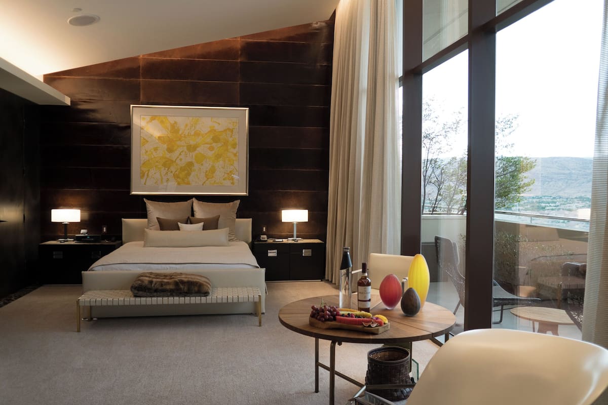 Een moderne hotelkamer met een groot bed, nachtkastjes met lampen, een ronde tafel met fruit en een fles, en een uitzicht op het omliggende landschap door grote ramen.