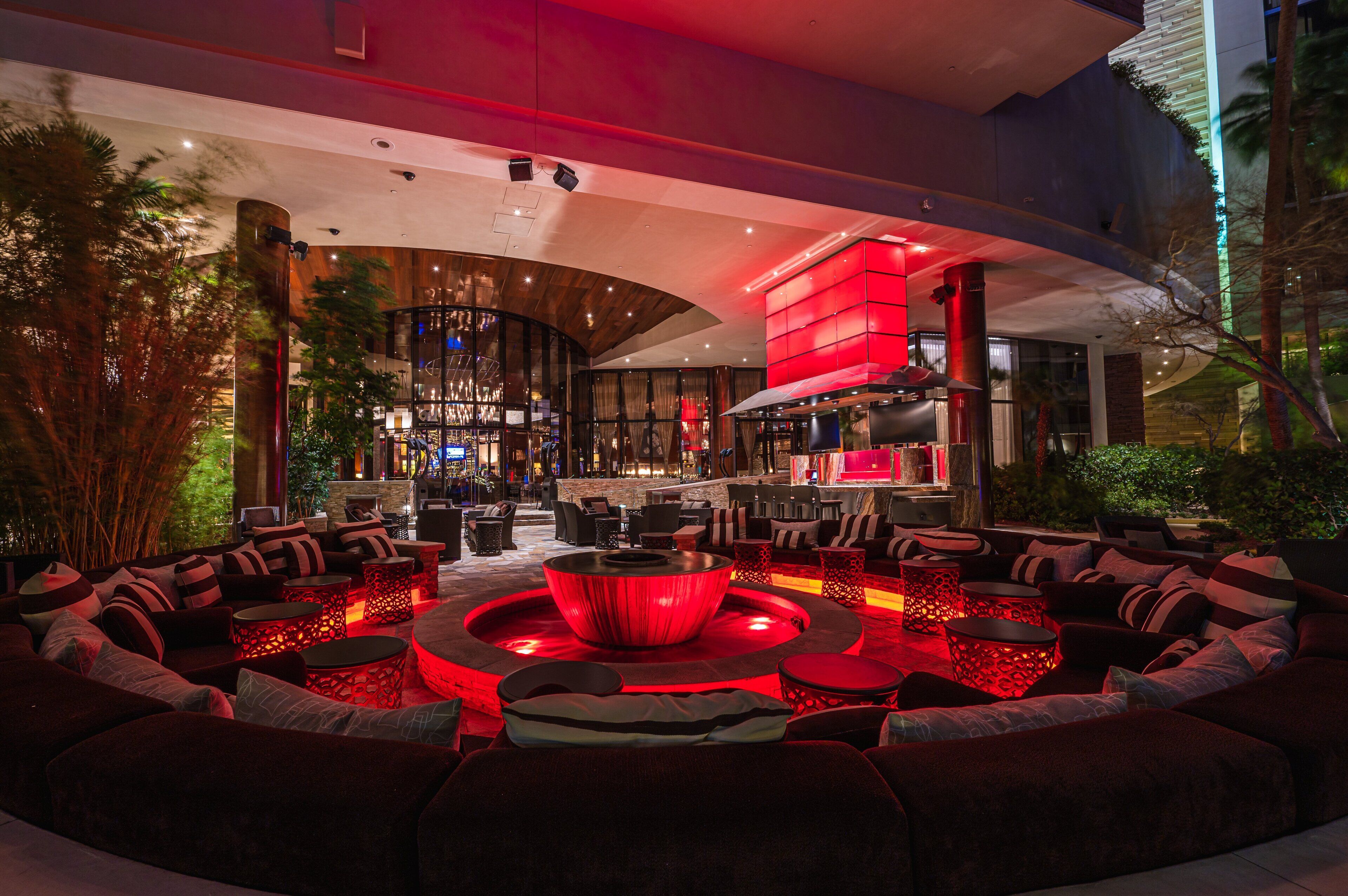 A modern lounge area with a circular seating arrangement, a central fire feature, and a bar area in the background.
