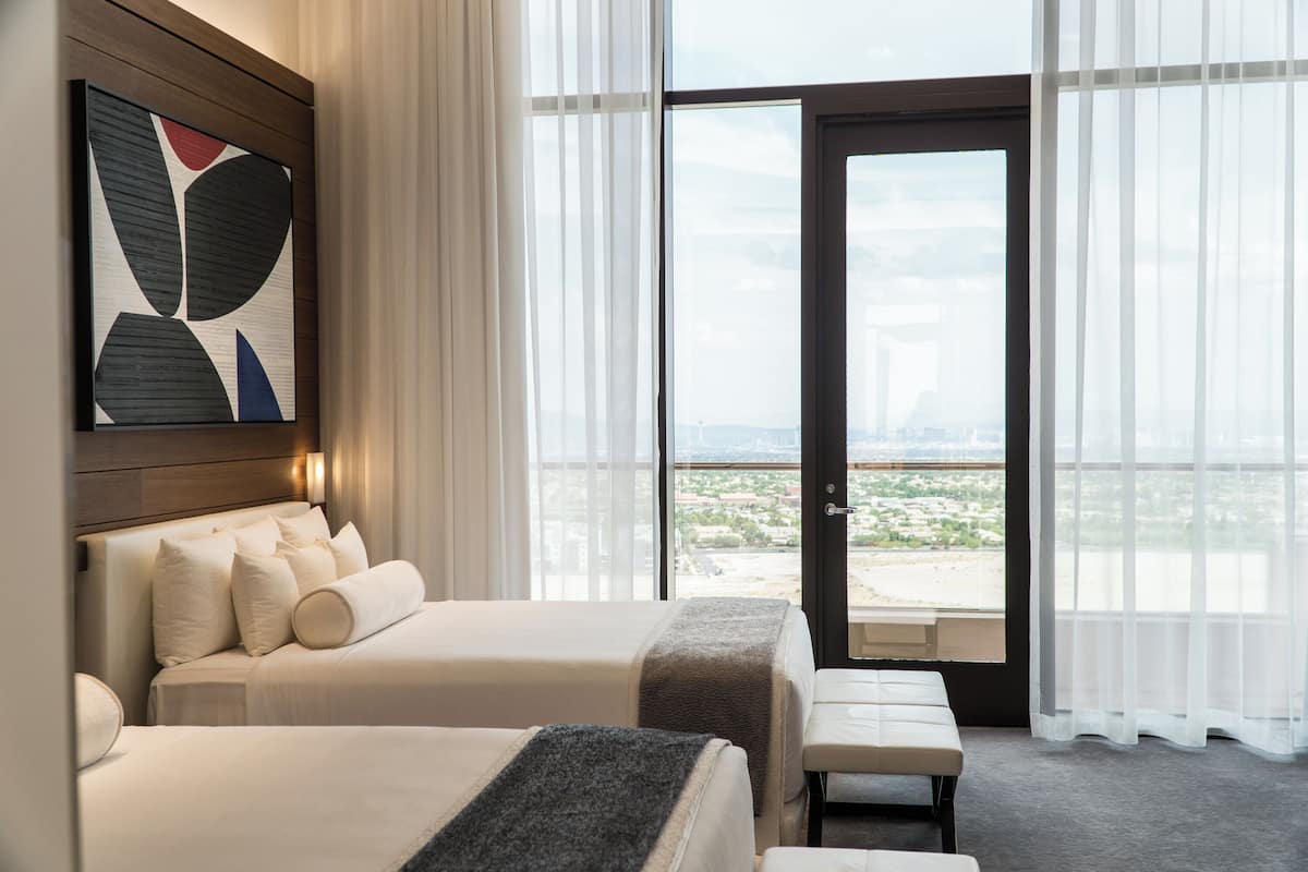 Een hotelkamer met twee bedden, een bankje, een groot raam met uitzicht op de stad en een deur naar buiten.