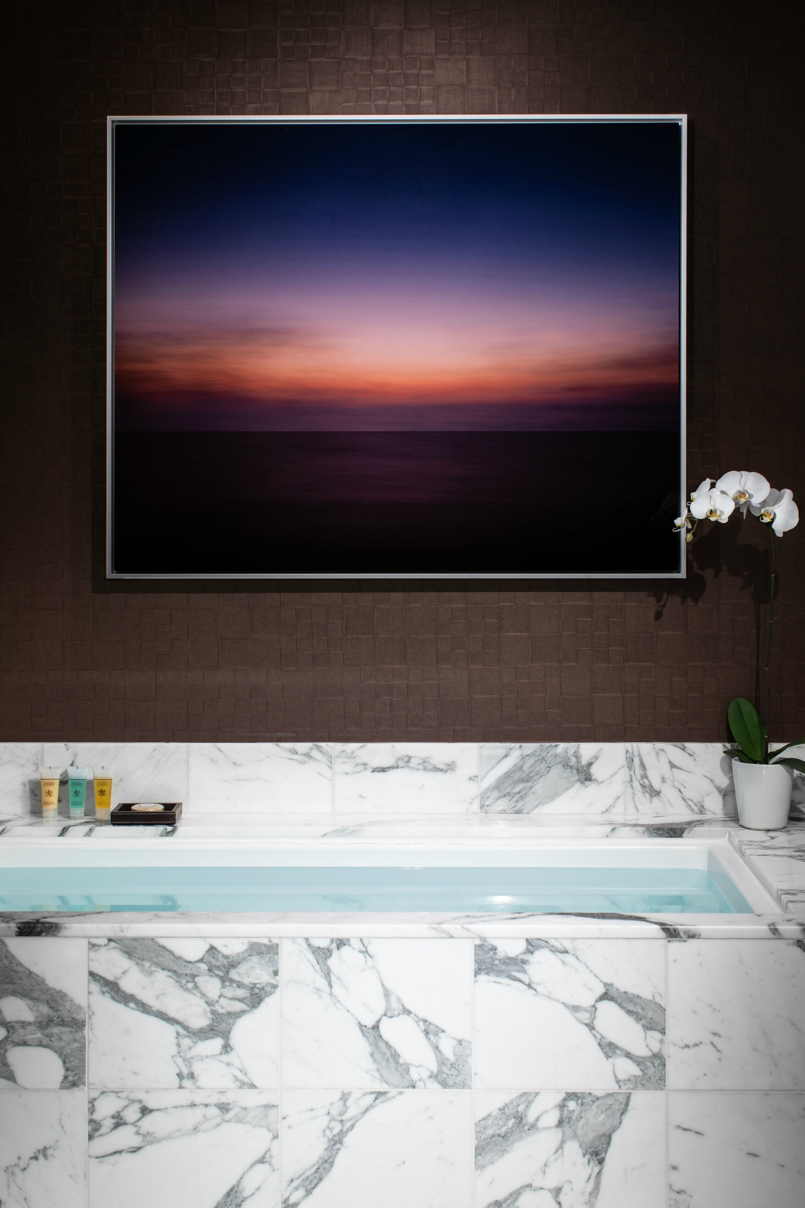 A bathroom with a marble sink, a large framed picture of a sunset, and a potted orchid.