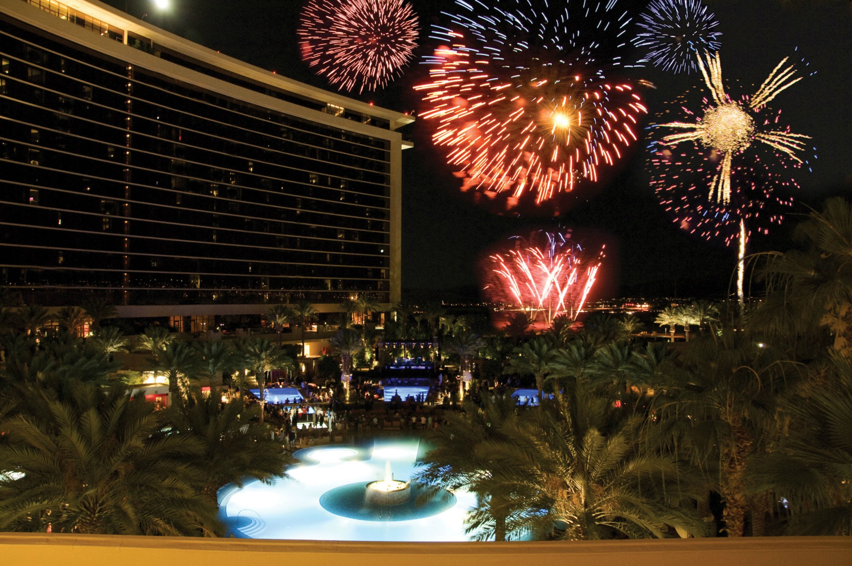 A hotel with a large building, a pool with a fountain, and fireworks in the background.