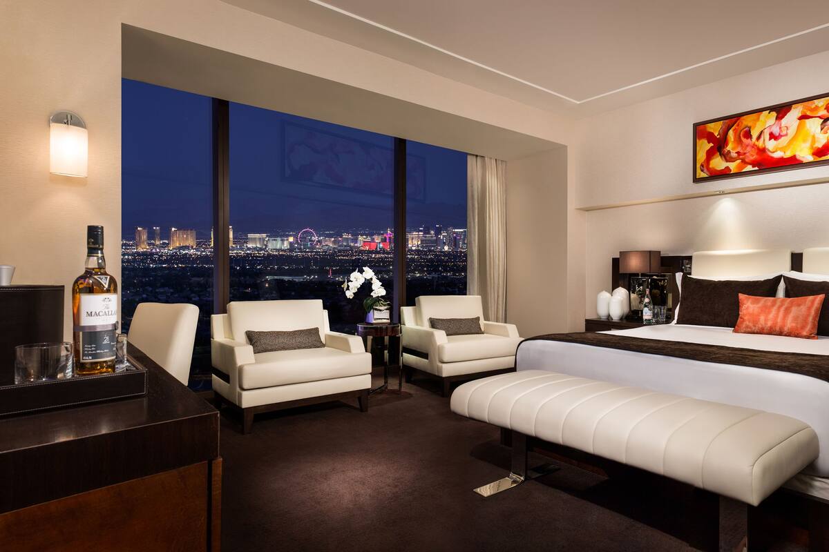 Hotelkamer met een groot raam met uitzicht op de stad, een bed met wit bedlinnen en een oranje kussen, een bankje en een nachtkastje met een lamp en decoratieve objecten.