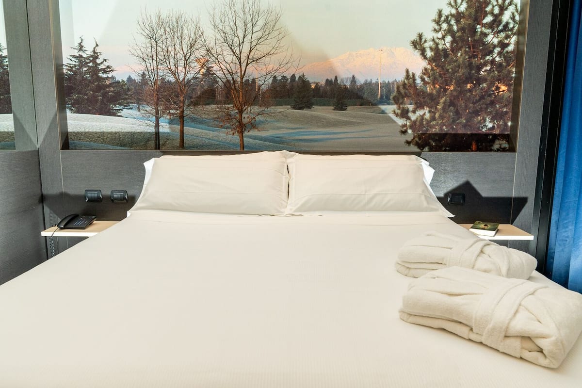 Habitación de hotel con una cama grande, una mesita de noche con teléfono y vistas a un paisaje nevado con árboles y una montaña.