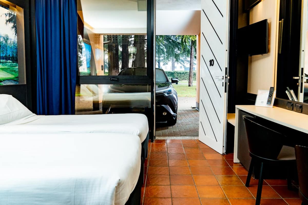 Habitación de hotel con cama, escritorio, televisor y vistas al exterior de un coche y árboles.