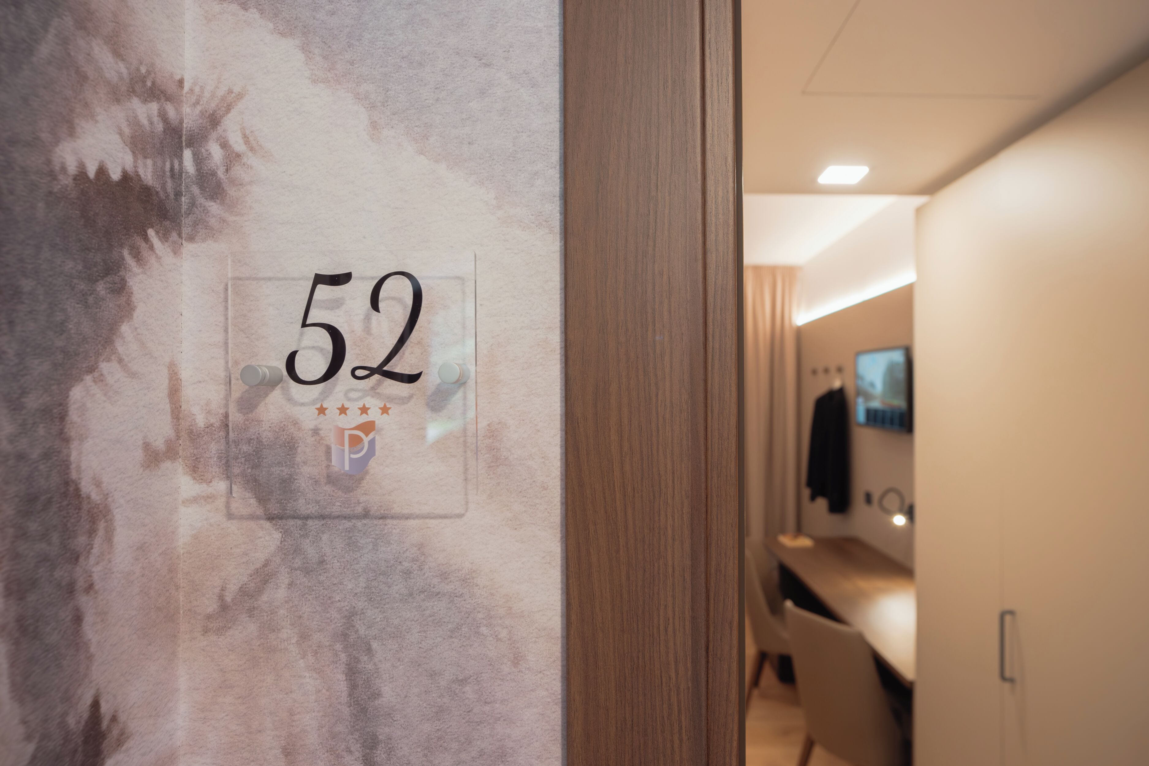A hotel room with a number 52 sign on the wall, a wooden door, and a glimpse of a modern bathroom with a mirror and sink.
