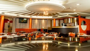 A modern hotel dining area with round tables, orange chairs, and a central decorative light fixture.