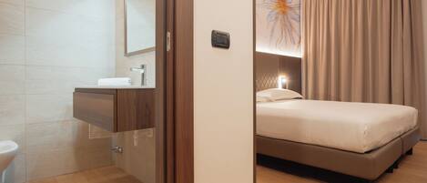 Una habitación de hotel moderna con piso de madera, una cama con cabecera, un baño con lavamanos y espejo, y un ventanal con cortinas.