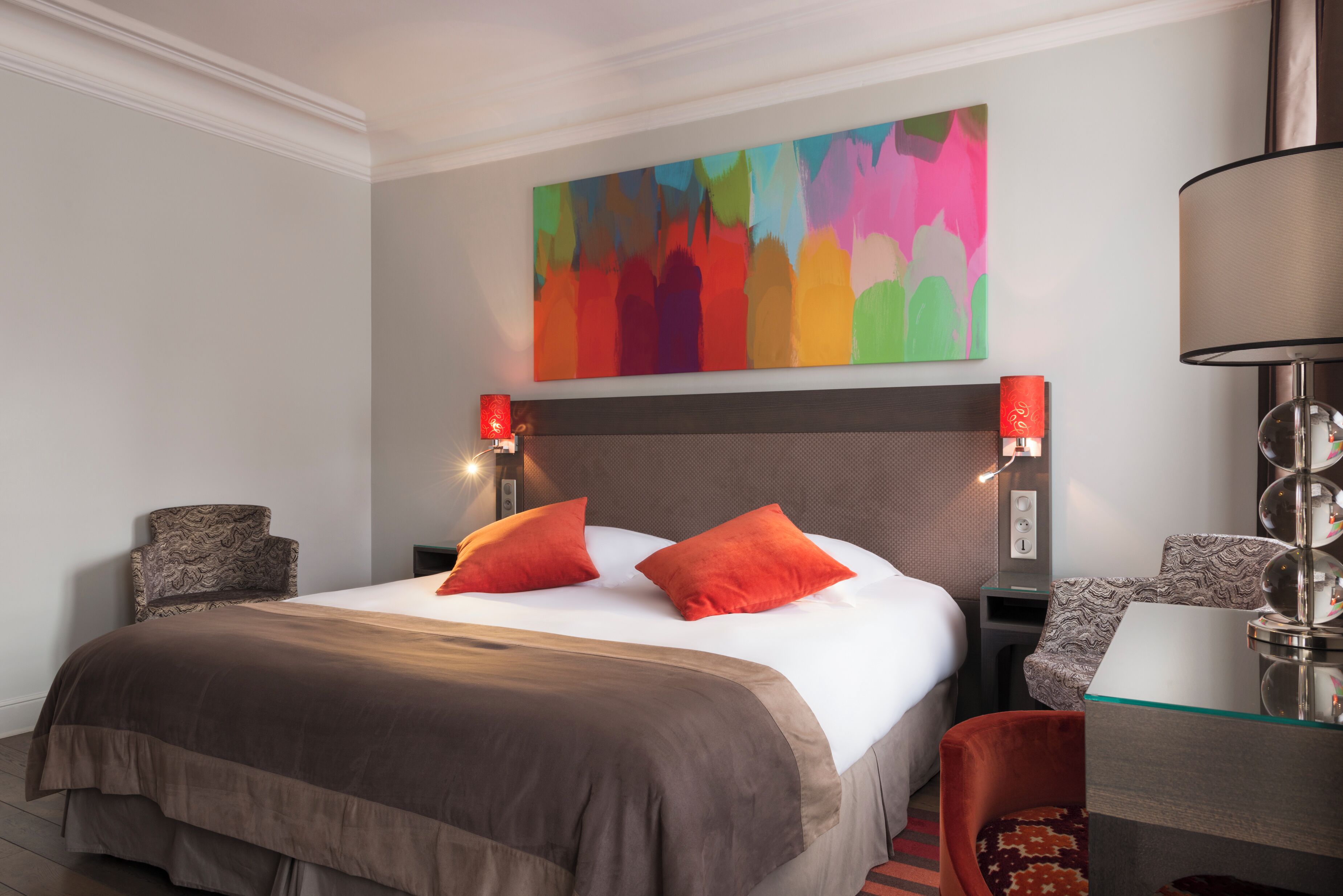 A hotel room with a large bed, two red pillows, a colorful abstract painting above the bed, a desk with a lamp, and a chair.