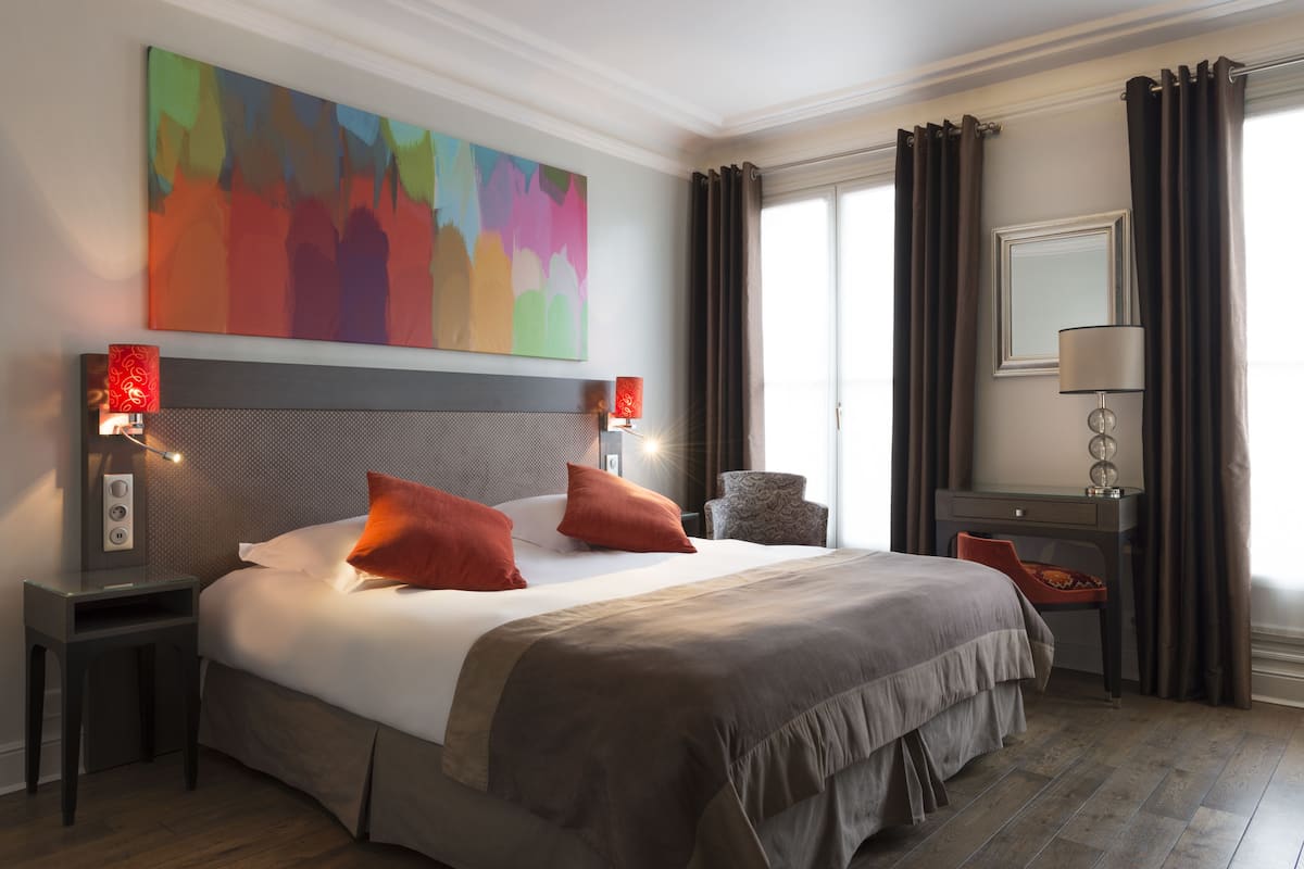 A modern hotel room with a large bed, two bedside tables with lamps, a desk, a chair, and a colorful abstract painting on the wall.