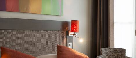 A hotel room with a large bed, two red pillows, a headboard with a textured pattern, a wall-mounted lamp, and a patterned armchair.