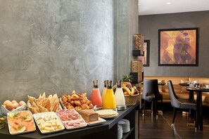 A breakfast buffet with various food items and beverages, a seating area with a couch and chairs, and a modern wall with abstract art. - Hôtel Régence Etoile (Paris)